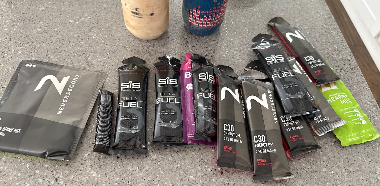 Cycling fueling products on a kitchen counter: a drink mix packet, SIS Beta Fuel gels, and Neversecond C30 energy gels ready for a long ride