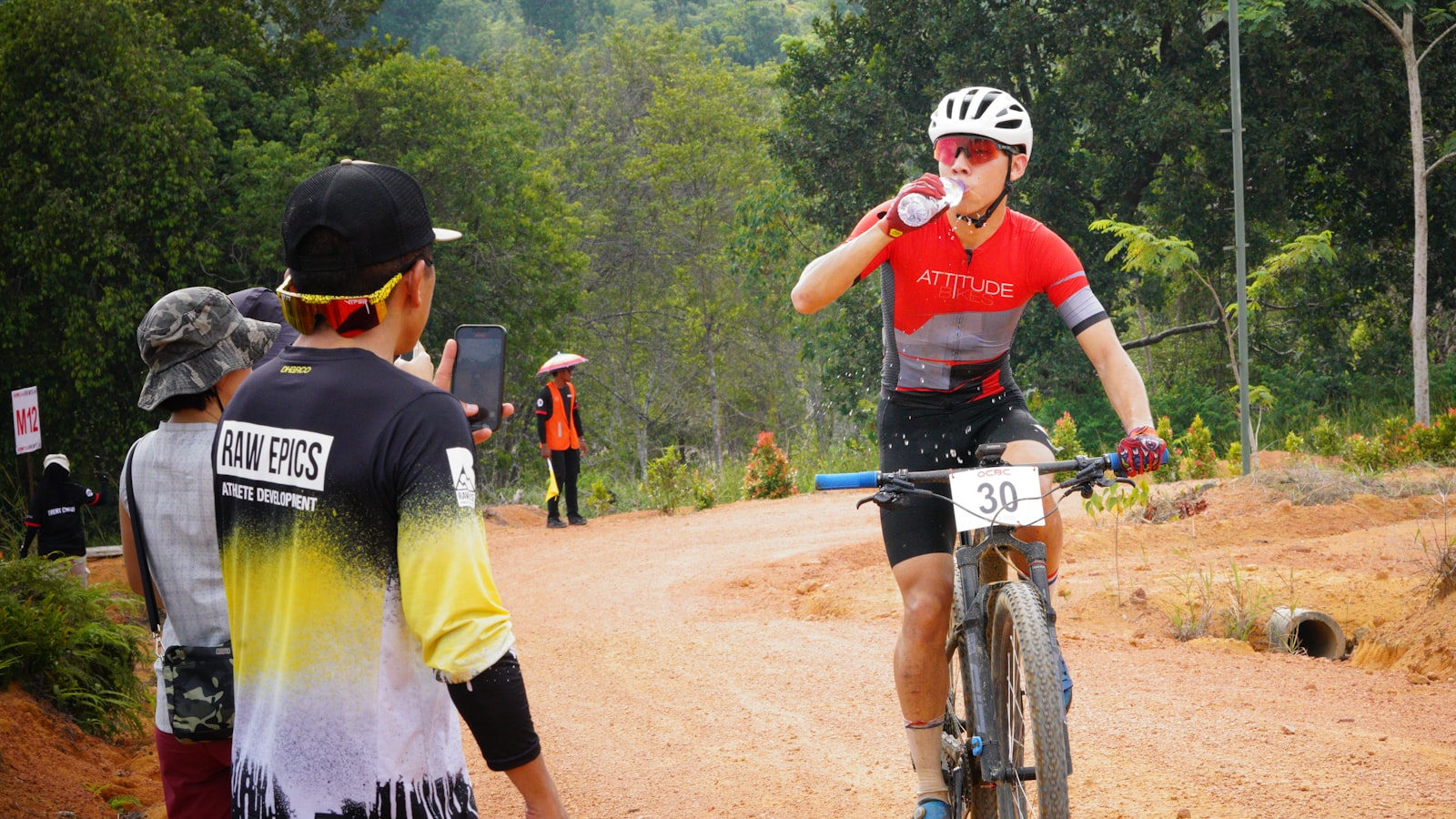 Mountain bike racer drinking from a water bottle mid-race on a dirt course, illustrating cycling hydration