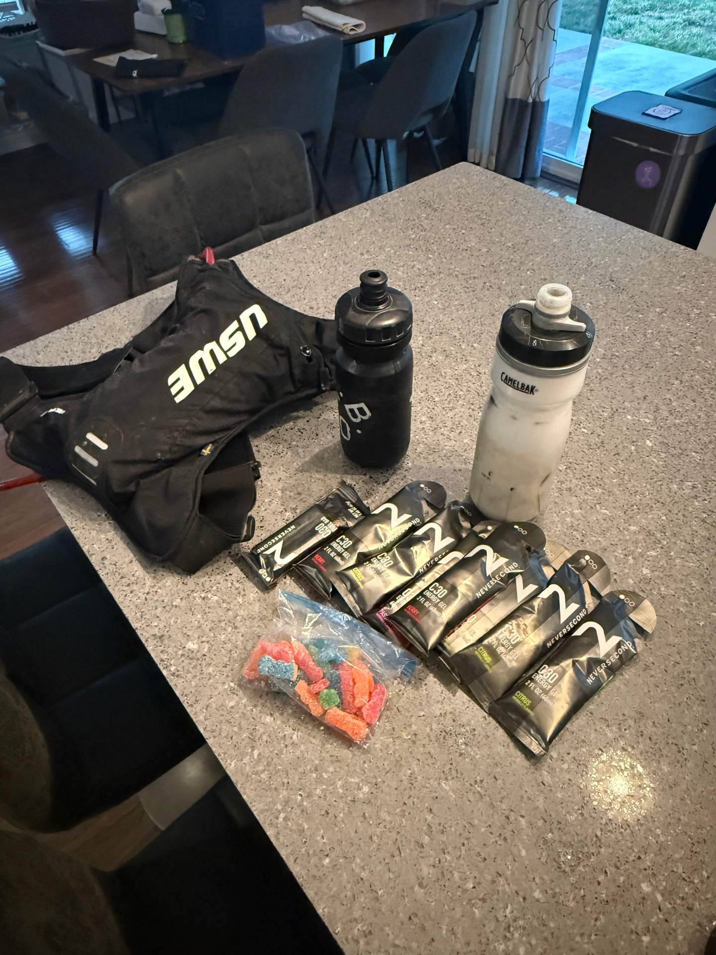 Race day fueling setup on a kitchen counter: USWE hip pack, CamelBak bottle, energy gels, and candy ready for a long ride